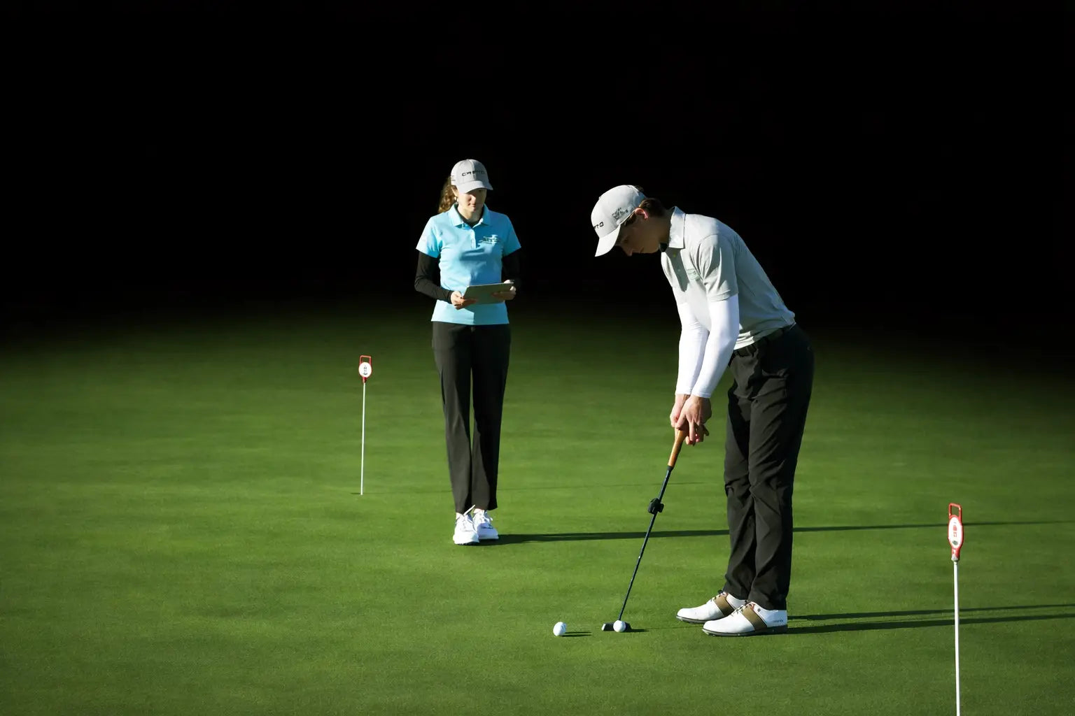 Two golfers on a putting green using a Capto putting analysis device, with one player preparing to putt while the other observes and records data.