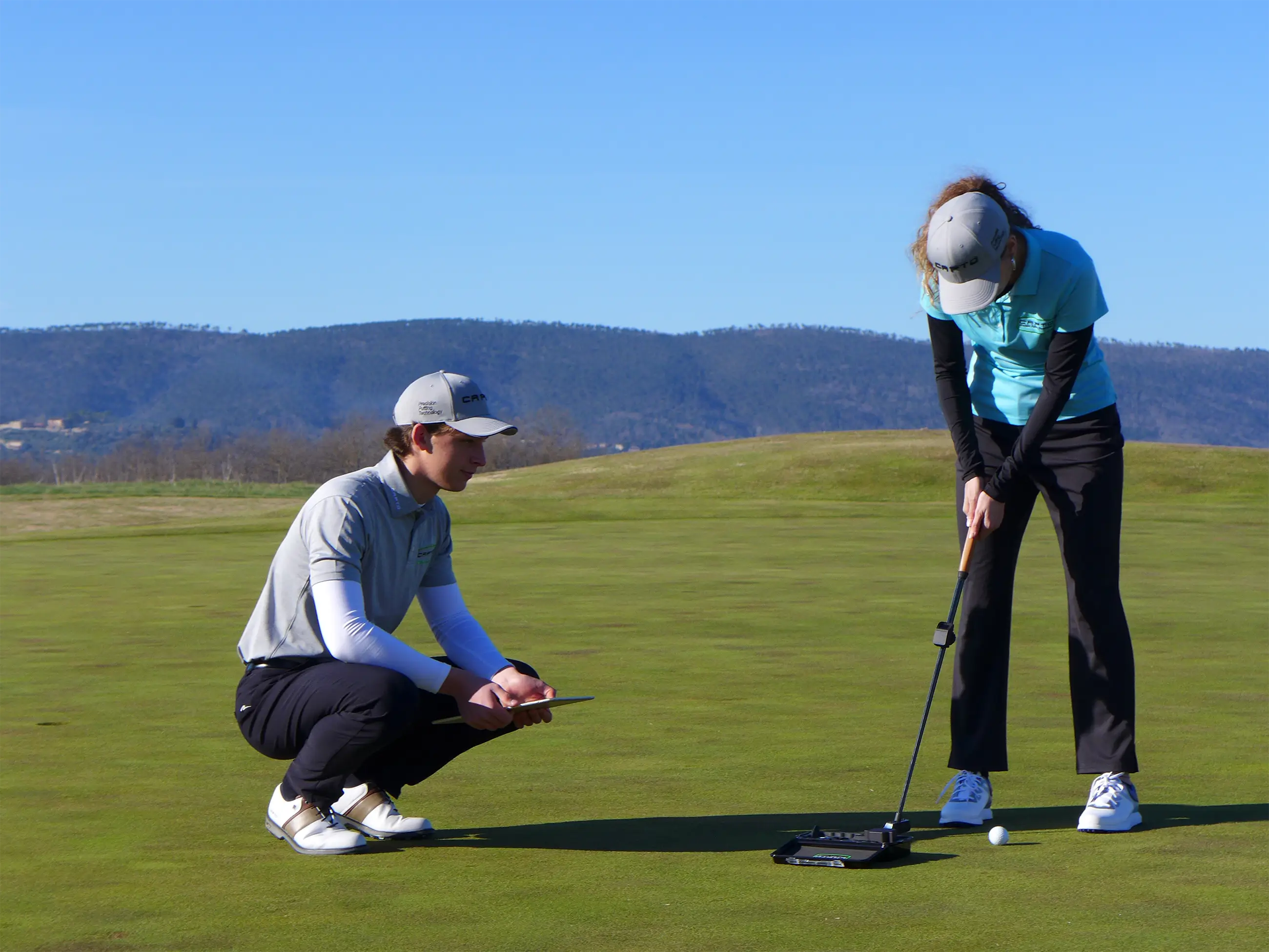 Two golfers on a putting green with a dark, softly faded background; a woman in a light blue polo focuses on her putt while a man crouches nearby holding a tablet, observing her technique.