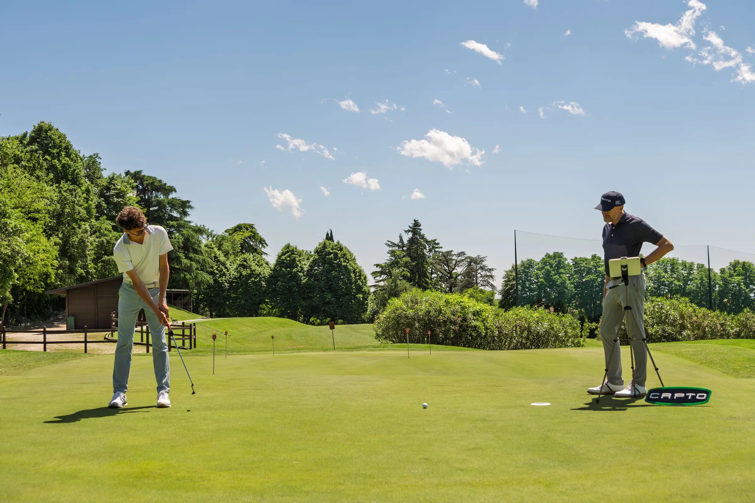 Two men on a golf putting green under a blue sky; one is putting while the other stands near a tripod with a tablet and a CAPTO device.
